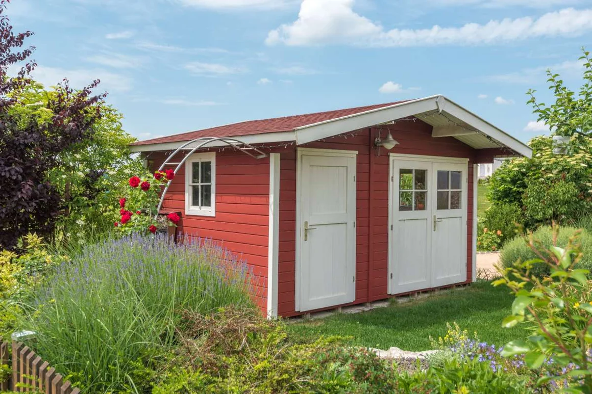 Ein rotes Gartenhaus mit weißen Türen und Fenstern in einem blühenden Garten.